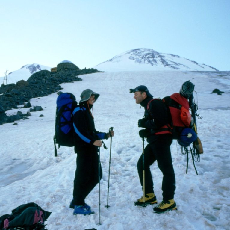 Zwei Bergsteiger mit Rucksäcken stehen im Schnee vor einem Gipfel.