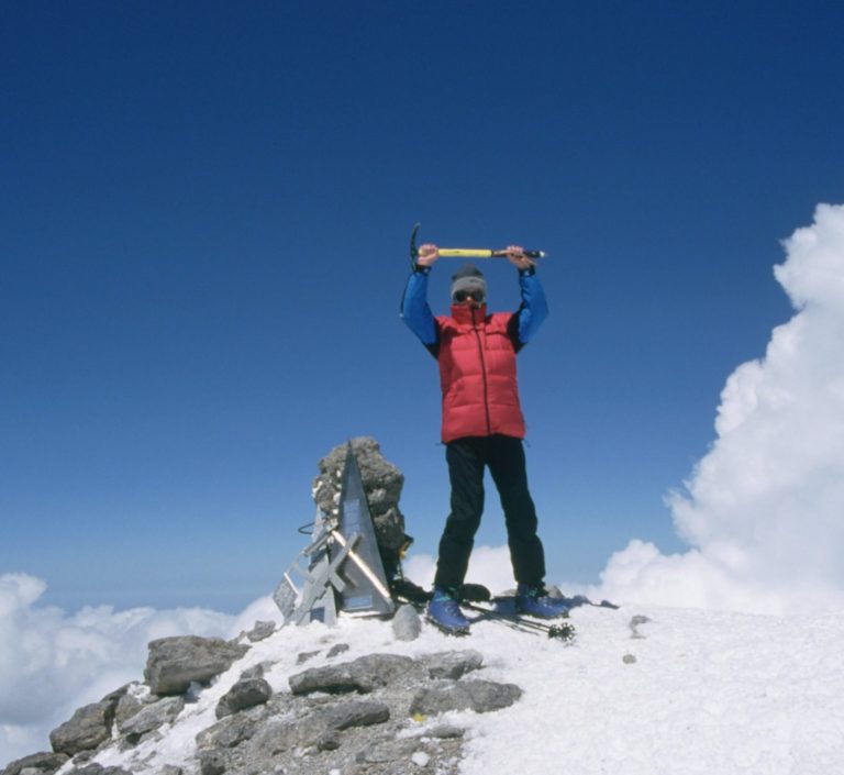 Person in rot-blauer Kleidung steht auf einem Gipfel und hält ein Schild hoch. Schnee und Wolken.